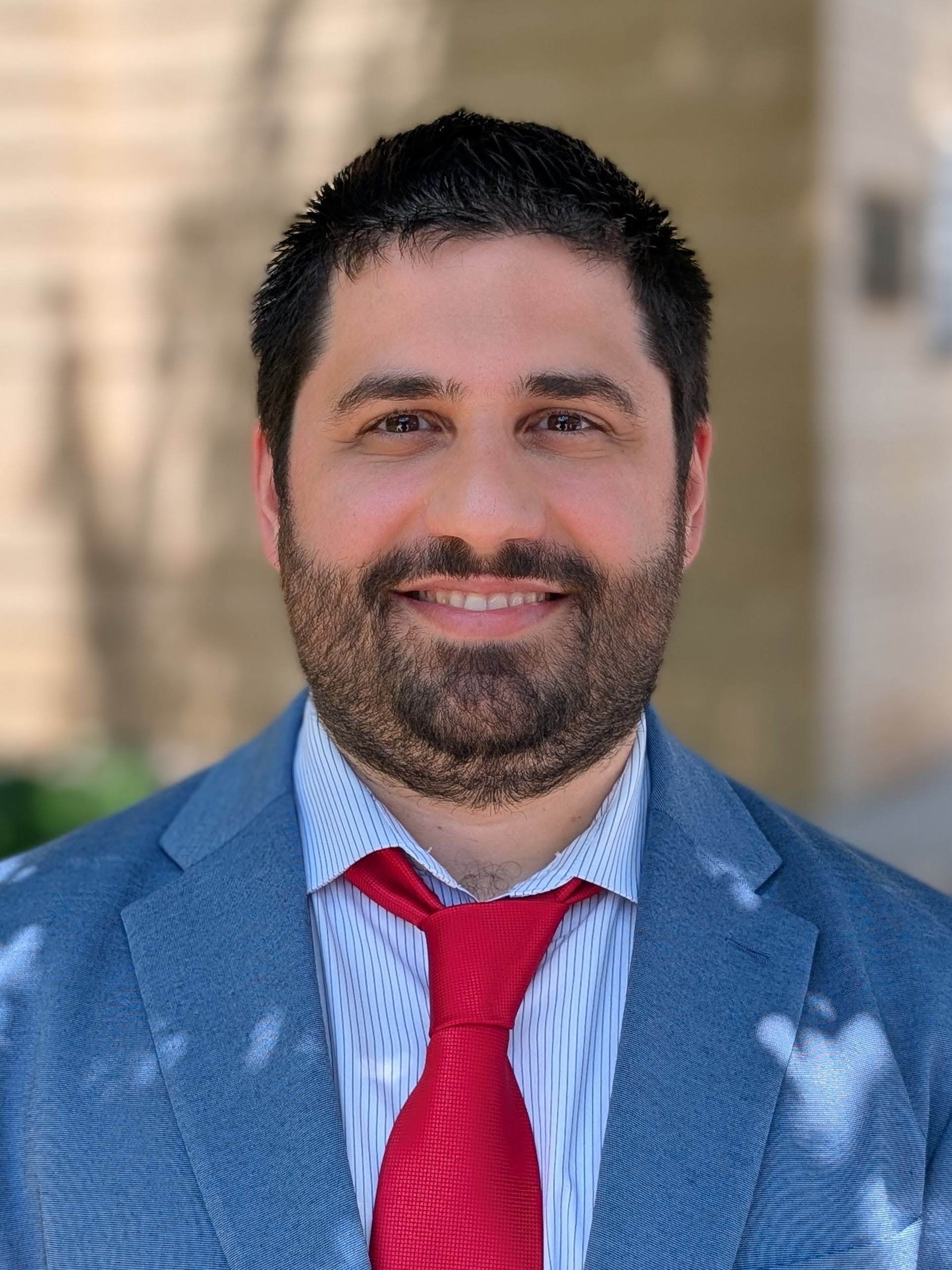 A portrait of João M Pereira, a handsome man in his mid 30s with a friendly smile, wearing a white shirt, UGA red tie and blue blazer.